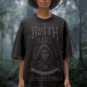 Könnte beinhalten: Ein schwarzes Kurzarm-T-Shirt mit einem Grafikdruck, der den Text Truth or Dare, Do you dare to know the truth? und With the Suriel zeigt. Das Design enthält eine Kapuzenfigur innerhalb eines Rahmens mit Sternakzenten.