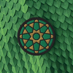 May include: A circular embroidered patch featuring a geometric star pattern in shades of gold and green. The design consists of a central green circle surrounded by an eight-pointed gold star, enclosed within a dark border on a green scale background.