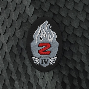 Op de afbeelding: Een ovaalvormige geborduurde patch met een gestileerd grijs vlamontwerp aan de bovenkant, een vetgedrukt rood cijfer 2 in het midden en een klein grijs schild aan de onderkant met het Romeinse cijfer IV, geflankeerd door twee kleine grijze vleugels.