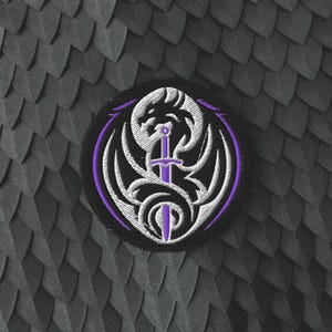 May include: A circular embroidered patch featuring a black background with a central purple sword design. The sword is surrounded by white, curved, wing-like shapes and a thin purple circular border, creating a detailed graphic emblem.