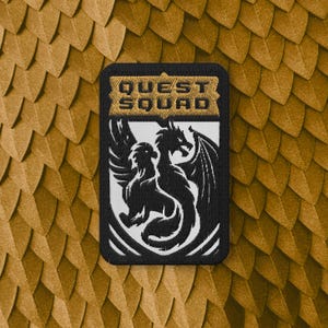 Puede incluir: Un parche rectangular bordado con un borde negro. La sección superior presenta el texto QUEST SQUAD en negro sobre un fondo dorado. La sección inferior muestra la silueta negra de un dragón sobre un fondo blanco.