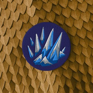 May include: A circular embroidered patch featuring a dark blue background with a central design of jagged, crystalline mountain peaks rendered in shades of light blue, white, and dark blue thread, creating a sharp, geometric, and textured appearance.