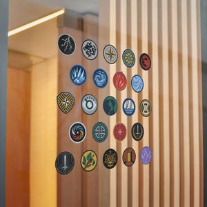 May include: A collection of twenty-four circular stickers arranged in a grid on a glass surface. Each sticker features a unique graphic design, including symbols like lightning, a compass, a shield, a sword, a tree, and various abstract patterns.