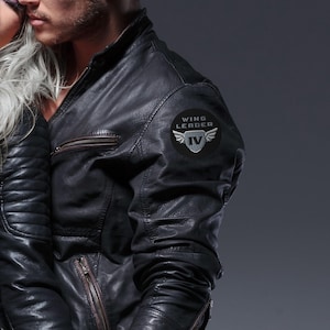 Könnte beinhalten: Eine schwarze Lederjacke mit einem runden Aufnäher am Oberarm. Der Aufnäher zeigt den Text WING LEADER über einem Schildsymbol mit Flügeln und der römischen Ziffer IV. Die Jacke verfügt über einen Stehkragen und Taschen mit Reißverschlüssen.
