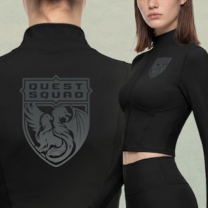 May include: A black long-sleeve zip-up jacket featuring a high collar and a cropped hem. The back displays a large gray shield emblem with a dragon graphic and the text "QUEST SQUAD," while a smaller matching emblem is printed on the chest.