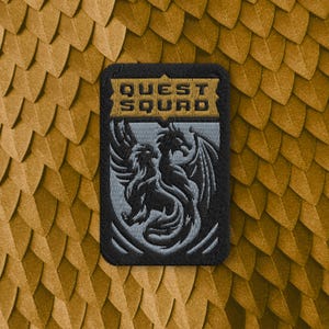 May include: A rectangular embroidered patch featuring a black border and a light grey dragon illustration centred on a dark background. The text QUEST SQUAD is stitched in gold thread across the top section within a banner-like design element.