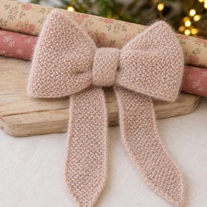 Könnte beinhalten: Ein weiches, hellrosa gestricktes Schleifenaccessoire mit einer texturierten kraus rechts Struktur. Die Schleife hat ein zentrales Knotendetail und zwei lange, spitz zulaufende Enden, gefertigt aus flauschigem Garn mit einem dezenten Glanz.
