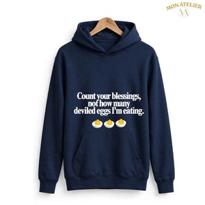 Peut inclure: Un sweat à capuche bleu marine avec le texte blanc, Count your blessings, not how many deviled eggs I'm eating, placé au-dessus d'une illustration graphique de trois moitiés d'œufs farcis avec des jaunes d'œufs jaunes.