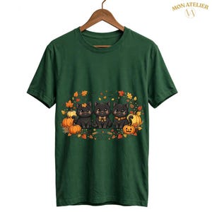 Peut inclure: Un t-shirt à manches courtes vert foncé avec un motif représentant trois chatons noirs assis parmi des citrouilles orange et des feuilles d'automne. Le texte MON ATELIER est imprimé dans le coin supérieur droit du t-shirt.