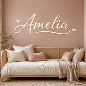 Può includere: Una scritta in legno da parete che riporta il nome Amelia in carattere corsivo, affiancata da due piccole stelle a cinque punte. Le lettere sono di colore crema chiaro, montate su una parete rosa tenue sopra un divano letto in rattan.