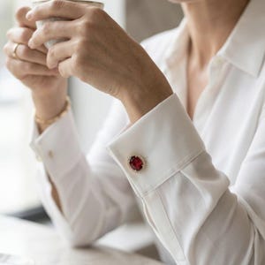 May include: A white long-sleeved button-down shirt featuring a prominent oval-shaped cufflink on the wrist. The cufflink showcases a deep red gemstone surrounded by a delicate border of small, clear, sparkling stones set in gold.