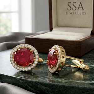Puede incluir: Un par de gemelos dorados con una piedra preciosa redonda de color rojo intenso en el centro, rodeada por un halo de pequeñas piedras blancas. Los gemelos se muestran frente a una caja de madera abierta con el texto SSA JEWELLERS impreso.