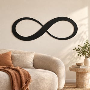 Puede incluir: Una pieza de arte mural de metal negro mate con forma de símbolo de infinito continuo. El diseño presenta un contorno suave y curvo con un centro abierto, creando una silueta audaz montada horizontalmente sobre una pared clara.