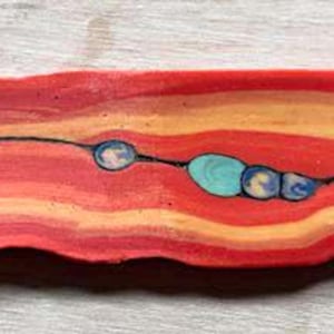 May include: A rectangular clay bar featuring horizontal wavy layers of red, orange, and yellow. A thin, dark, horizontal fissure runs through the center, punctuated by small, embedded circular and oval accents in shades of turquoise and blue.