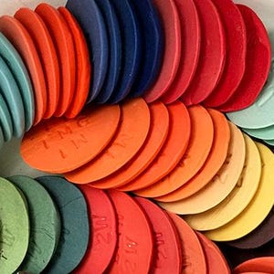 May include: A collection of circular, flat discs arranged in a curved formation, featuring a wide spectrum of colors including red, orange, yellow, green, blue, and purple. Each disc has embossed alphanumeric text markings on its surface.