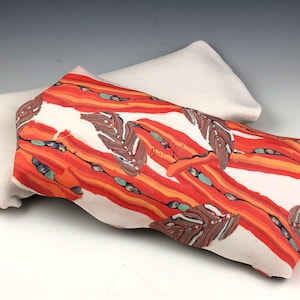 Peut inclure: Une forme d'oreiller en céramique rectangulaire avec une base blanche. La surface supérieure présente un motif vibrant de rayures diagonales orange et rouges, entremêlées d'illustrations de plumes brunes et de petits accents turquoise.