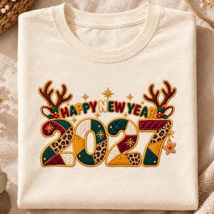 May include: A cream-colored crew neck t-shirt featuring an embroidered graphic that reads HAPPY NEW YEAR 2027. The text is accented with brown reindeer antlers, holly berries, and stars. The numbers are filled with patchwork patterns.