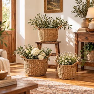 Op de afbeelding: Drie ronde gevlochten zeegrasmanden met handvatten aan de zijkanten, elk gevuld met groen of bloemen. Eén mand staat op een houten krukje, terwijl twee andere op de vloer staan. De manden hebben een natuurlijke kleur en een gevlochten textuur.