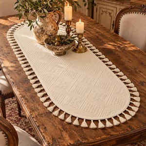 Puede incluir: Un camino de mesa ovalado de color crema con un patrón geométrico texturizado y un fleco decorativo de pequeñas borlas a lo largo de todo el perímetro. El camino está elaborado con un diseño tejido que muestra motivos cuadrados repetidos.