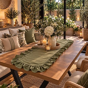 May include: A rectangular olive green table runner featuring a ruffled edge trim. The runner is laid flat across a wooden dining table, serving as a base for a ceramic pitcher filled with white flowers, a glass candle holder, and a small book.