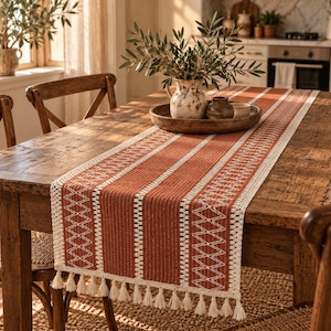 Può includere: Un runner da tavola rettangolare caratterizzato da strisce verticali alternate in tessuto color arancio bruciato e pizzo all'uncinetto color crema con un motivo geometrico a zigzag, rifinito con una frangia decorativa di nappe color crema.