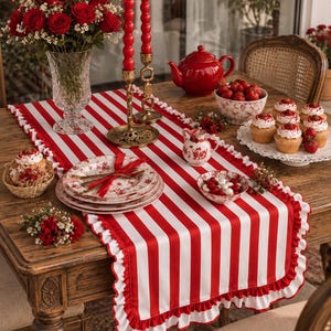 Puede incluir: Un camino de mesa rectangular con rayas verticales rojas y blancas y un borde con volantes. El camino de mesa se exhibe sobre una mesa de madera decorada con velas rojas, un jarrón de cristal con rosas rojas y vajilla con motivos florales.