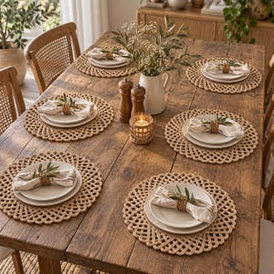 May include: A rustic wooden dining table set for six with round woven placemats, white ceramic plates, and linen napkins secured with rattan rings. A white pitcher with greenery, two wooden salt and pepper mills, and a small candle holder sit centre.