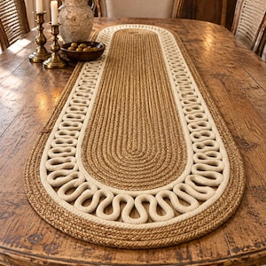 Peut inclure: Un chemin de table ovale fabriqué en corde de jute tressée de couleur beige naturel, présentant une section centrale unie entourée d'une bordure décorative en corde épaisse crème disposée en un motif ondulé continu et répétitif.