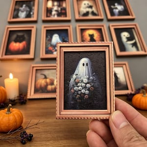 Può includere: Una cornice rettangolare in miniatura con finitura in rame metallizzato contenente l'immagine di un fantasma avvolto in un lenzuolo bianco che tiene un bouquet di fiori bianchi e arancioni su sfondo scuro, tenuta davanti a una parete di cornici simili.