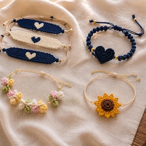 May include: A collection of five bracelets including three flat woven bands with heart motifs, one dark blue macrame bracelet with a central heart charm and gold beads, one floral crochet bracelet, and one sunflower crochet bracelet with a thin chain.