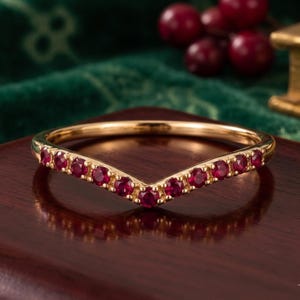 May include: A gold chevron-shaped ring featuring a row of small, round, deep red rubies set along the V-shaped band. The metal has a polished finish, and the stones are evenly spaced across the front curve of the ring, creating a symmetrical design.