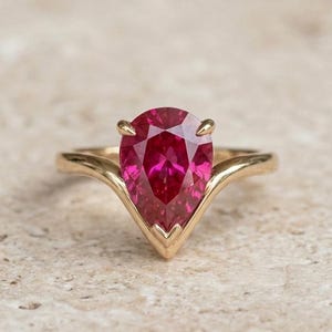 Puede incluir: Un anillo de oro con una piedra preciosa en forma de pera de color rojo rosado intenso en un engaste en forma de V. La banda es lisa y pulida, curvándose hacia arriba hasta la base puntiaguda de la piedra facetada, sujeta por tres garras metálicas.