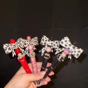 Op de afbeelding: Vier pennen met decoratieve kralen en stoffen strikken. Twee pennen hebben rode en roze glitterbehuizingen met luipaardprint strikken, terwijl de andere twee zwarte glitterbehuizingen hebben met wit-zwarte koeienvlekken strikken.