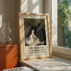 Könnte beinhalten: Ein gerahmter Kunstdruck mit einer schwarz-weißen Katze mit grünen Augen. Der Text lautet THE INDOOR CAT SOCIETY EST. SOMEWHERE BETWEEN NAP TIME & DINNER. Der Druck befindet sich in einem hellen Holzrahmen mit einem dekorativen Randdesign.