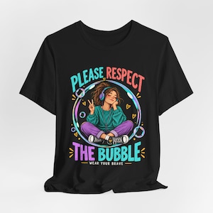 May include: A black t-shirt featuring a graphic of a person sitting cross-legged inside a large bubble, wearing headphones, and making a peace sign. The text reads PLEASE RESPECT THE BUBBLE and WEAR YOUR BRAVE in colourful, bold lettering.