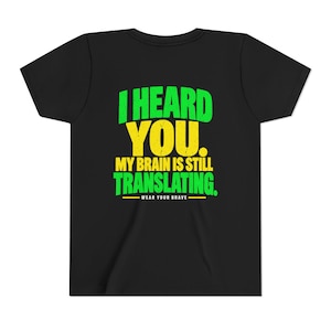 May include: A black short-sleeve t-shirt featuring the green and yellow text I HEARD YOU. MY BRAIN IS STILL TRANSLATING. and the smaller white text WEAR YOUR BRAVE with a yellow line underneath, centered on the front of the garment.