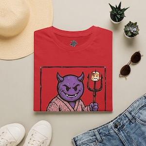 Puede incluir: Una camiseta roja de cuello redondo con un estampado gráfico de un personaje de diablo morado que lleva un suéter de punto rosa. El personaje sostiene un tridente negro con una nube tostada en la punta. El diseño está dentro de un borde negro.
