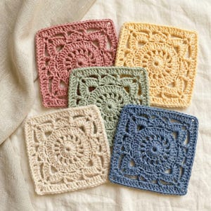 May include: Five square crochet coasters in dusty pink, pale yellow, sage green, cream, and slate blue. Each coaster features a circular floral pattern worked into a square shape with intricate openwork stitches and a scalloped border edge.