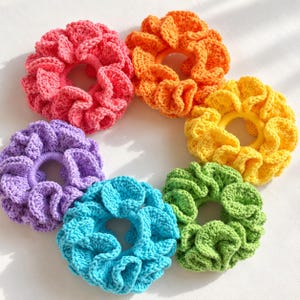 May include: A collection of six circular crochet hair scrunchies arranged in a semi-circle. The set includes individual scrunchies in vibrant shades of pink, orange, yellow, green, blue, and purple, each featuring a ruffled, textured yarn design.