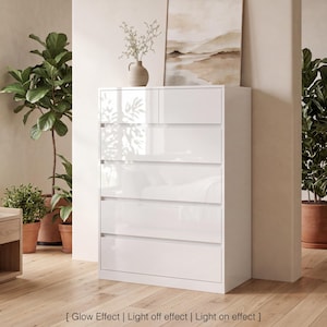 May include: A tall white chest of drawers with a high-gloss finish, featuring one small top drawer and four larger drawers below. The unit has a clean, rectangular design with integrated handles and a smooth, reflective surface throughout.