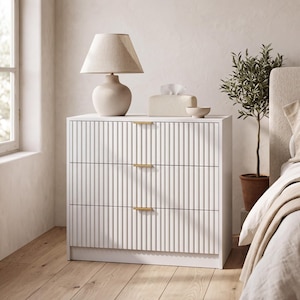 May include: A white three-drawer dresser featuring a vertical fluted texture on the drawer fronts and slim, horizontal gold-toned metal handles. The dresser has a rectangular frame and sits on a flat base, providing storage in a bedroom setting.