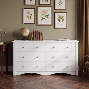 May include: A white six-drawer dresser featuring a rectangular frame with a curved bottom edge and small, round, silver-toned knobs on each drawer. The dresser has a smooth, matte finish and is positioned horizontally against a wall.