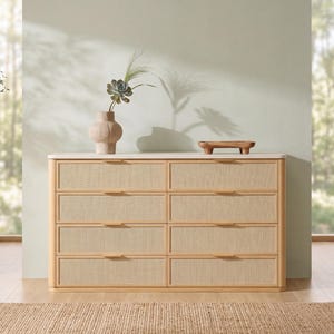 May include: A light wood dresser featuring eight drawers with woven cane panels and slim horizontal handles. The top surface is smooth and white, holding a textured beige vase with a succulent and a small wooden tray with four short legs.