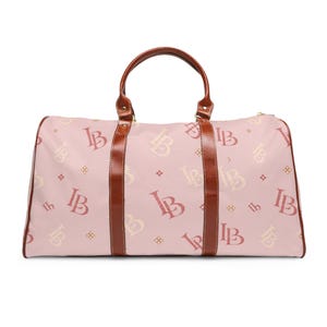 May include: A light pink duffel bag featuring a repeating pattern of the letters LB in dark red and cream. The bag includes two brown leather-like handles and brown vertical trim accents that extend from the top handles to the bottom base.