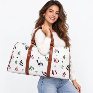 May include: A white duffel bag featuring a repeating pattern of the letters IB in black, dark red, and dark green. The bag includes brown leather-like handles and trim, with small four-petal flower accents scattered throughout the design.
