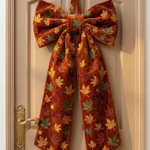 May include: A large, burnt orange velvet bow hanging on a door, featuring an embroidered pattern of scattered maple leaves in shades of yellow, green, and deep red. The bow has two long, trailing ribbons that extend downward from the center knot.