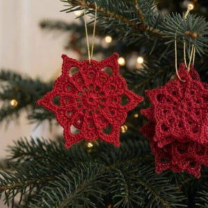 Puede incluir: Un conjunto de adornos de ganchillo de color rojo intenso en forma de estrella con intrincados patrones calados. Cada adorno está hecho de hilo y cuelga de un fino cordón metálico dorado, diseñado como pieza decorativa colgante.