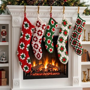 Op de afbeelding: Vijf handgemaakte gehaakte kerstsokken hangend aan een witte schoorsteenmantel. De sokken hebben een klassiek granny square patroon in afwisselende kleuren rood, wit en groen garen, opgehangen aan goudkleurige metalen haken voor kerstsokken.
