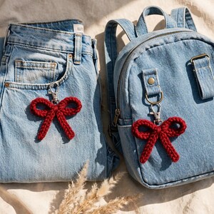 May include: Two dark red crocheted bow keychains with silver-toned metal lobster clasps. One keychain is attached to the pocket of light blue denim jeans, and the other is clipped to the front strap of a small light blue denim backpack.