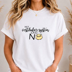 Könnte beinhalten: Ein weißes Kurzarm-T-Shirt mit dem schwarzen Text "MY vestibular system SAID NO" und einer kleinen, runden, gelben Cartoon-Figur mit großen Augen und Bewegungslinien, die Schwindel andeuten, neben dem Wort NO.